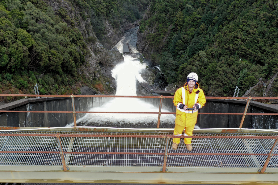 Oliver, wearing high-vis and a helmet, smiling for a photo standing in front of a a dam spilling water downstream. A wall of green trees is in the background.