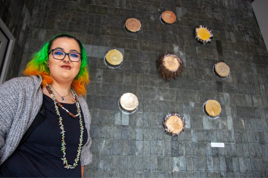 Artist Bianca Templar smiling, standing in front of her new artwork "From Coast to Country". The artwork is made up of 9 circular disks that includes weaving, ochre, shells, feathers and resin, formed in the shape of a petroglyph.