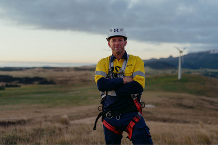 A person in high-vis workwear and a hardhat, looking at the camera. Behind him is an open field with a wind turbine in the background, and a cloudy sky overhead.