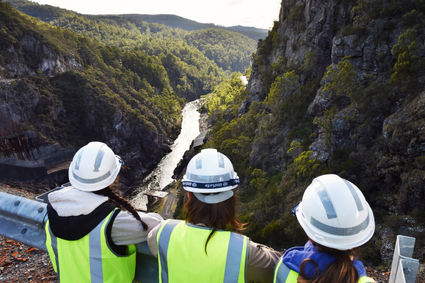 The back of three girls with high-vis and hard hats, on a dam wall looking downstream. The water flows through a valley of cliff-face and tree linings.
