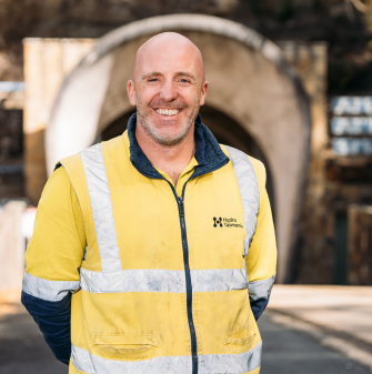 Craig is wearing a high-vis jacket and smiling towards the camera. He is standing in front of Poatina Power Station tunnel.