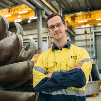 Xavier is wearing a high-vis shirt, and smiling for the camera. On his left is a large turbine part, and behind him is a workshop interior.