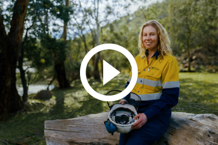 Leah Powell, a Hydro Tasmania employee, in high-vis clothing and holding a helmet. Leah is smiling for the photograph whilst sitting on a log, with green grass and trees behind her.